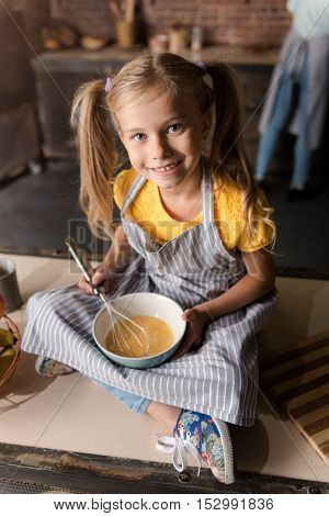 In a cheerful mood. Delighted amused smiling girl holding the bowl and mixing eggs while sitting on the table in the kitchen and her mother cooking in the background