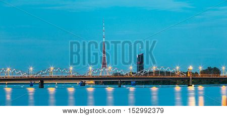 Riga, Latvia. Two Bridges Over Daugava Together: Stone Akmens And Railway Bridge In Evening Illumination With Red Radio TV Tower Background In Summer Dusk Under Blue Sky.