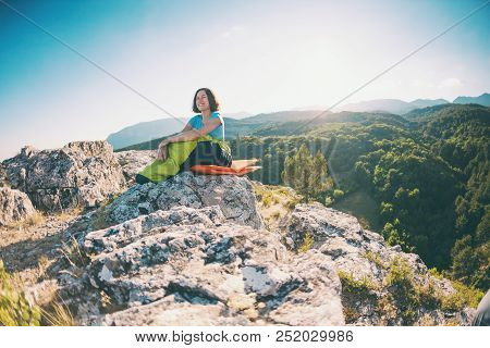 A Woman Is Sitting In A Sleeping Bag At Sunset. Camping On Top Of A Mountain. A Smiling Girl Rests A