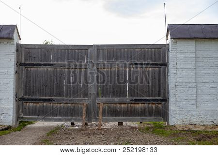 Old Wooden Gate With Dirt Road And Two Wooden Balks In A Gray Brick Fence.