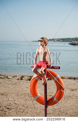 Cute Little Boy In Sunglasses Sits On Pole With Orange Lifebuoy Hanging On It On California Beach
