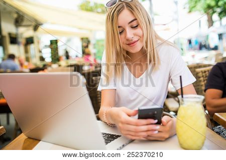 Thoughtful Young Woman Holds Phone, Uses Laptop Computer For Online Communication, Drink Cockail At