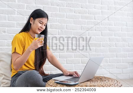 Asian Female Sit On Floor Using Laptop On Wicker Stand And Drink Orange Juice In Living Room At Home