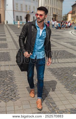 sexy young man wearing sunglasses and leather jacket walking in the city and looking to side, full body picture