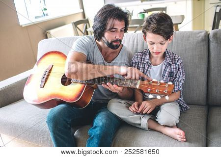 Man Is Istting On Sofa With His Son. He Holds Guitar. Man Is Pointing On Strings. Boy Looks At It . 