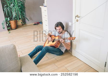 A Picture Of Gy Sitting Alone On The Floor And Playing The Guitar. He Is Leaning To The Wall. Man Is