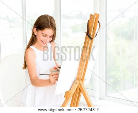 Pretty girl with brush in hand. Creative teen girl paitning a picture on easel. Girl practising drawing in art school
