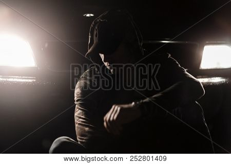 Fashionable Young Model Man In A Black Cap In Stylish Clothes With A Hooded Sits Near A Car With Lig