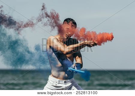 Attractive Shirtless Man Dancing With Red And Blue Smoke Sticks In Front Of Ocean View