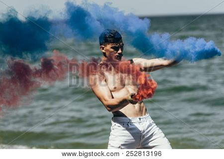 Shirtless Man Dancing With Red And Blue Smoke Sticks In Front Of Ocean View