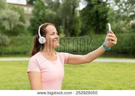 fitness, technology and healthy lifestyle concept - smiling woman in headphones listening to music and taking selfie by smartphone at park