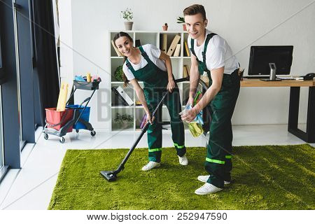 Happy Young Janitors Claning Carpet And Smiling At Camera In Office