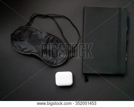 Flat Lay Of Black Notebook, Black Mask For Sleep And Wireless Headphones On Black Background