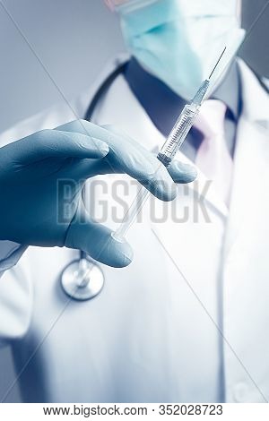 Doctor In Gauze Mask And Medical Robe Holding Syringe In His Blue Rubber Glove Dressed Hand. Health 