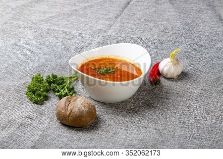 Beef Goulash Soup With Tongue. A White Oval Plate With Red Soup Stands On A Gray Canvas. Next To It 