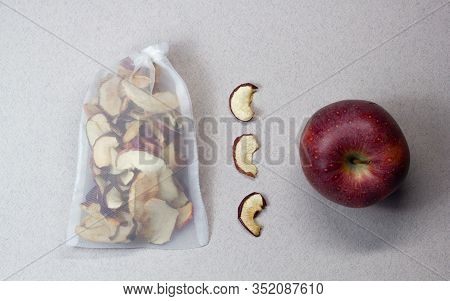Dried Apple Chips In A Transparent Cloth Bag, Three Slices Of Dried Apples And A Red Apple On A Gray