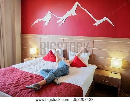 Sochi, Russia - 13 October 2019. A Girl Lies On A Bed In A Hotel Room With A Red Wall And White Moun