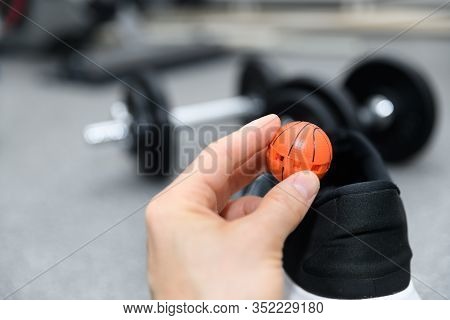 Hand Holding Shoe Freshener Ball In Front Of Athlete Footwear With Dumbells In The Background. Conce