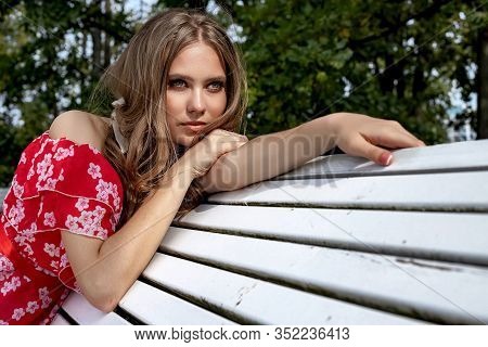 Beautiful Young Girl, Model, Brown-haired Woman With Long Hair In A Red Dress Sits On A Bench And Po