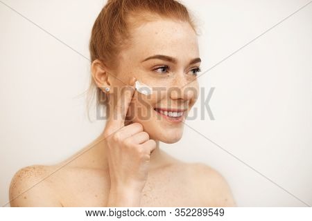Lovely Caucasian Girl With Red Hair And Freckles Is Smiling While Using A Cream On Her Face