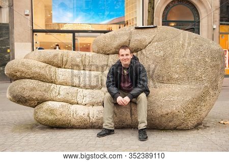 Young Caucasian Man Sitting On Huge Stone Hand On Street Of Antwerp, Belgium In Early Spring Day. An