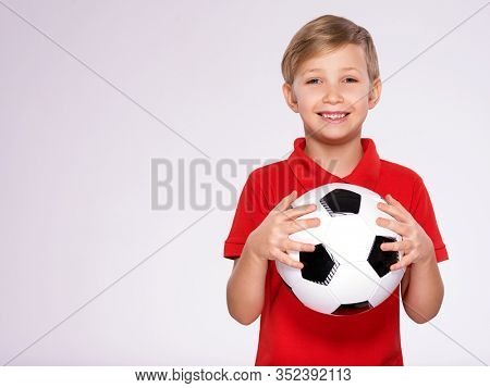 Photo of a smiling boy in sportswear holding soccer ball, posing at studio. Happy 8 years old kid in a red t-shirt with a soccer ball in hand. White child with a smile holds a soccer ball.