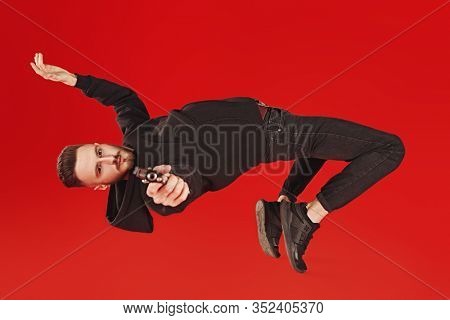 Handsome man in black clothes in motion in flight with a gun. Super agent on a mission. Criminal world, counterterrorism. Studio shot on a red background.