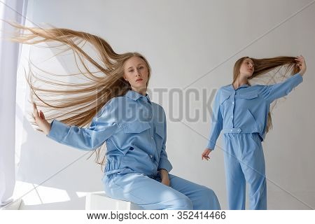Two Young Pretty Twin Sisters Posing In Blue Suits On White Studio Cyclorama. One Of Them Sits On Wo