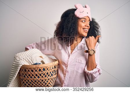 Young african american woman with afro hair wearing pajama doing laundry domestic chores pointing and showing with thumb up to the side with happy face smiling