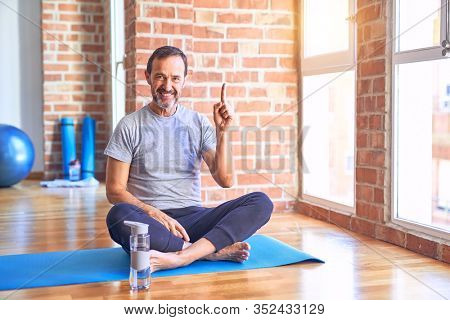 Middle age handsome sportman sitting on mat doing stretching yoga exercise at gym pointing finger up with successful idea. Exited and happy. Number one.