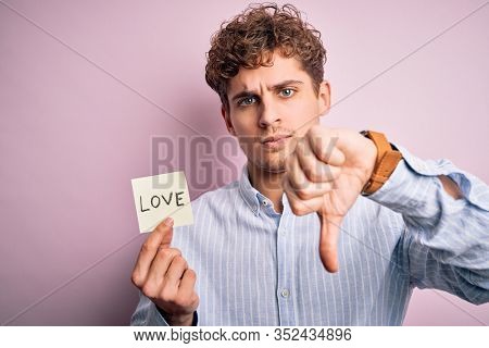 Young blond romantic man with curly hair holdiing paper with love message for valentines day with angry face, negative sign showing dislike with thumbs down, rejection concept