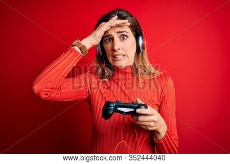 Young beautiful brunette gamer woman playing video game using joystic and headphones stressed with hand on head, shocked with shame and surprise face, angry and frustrated. Fear and upset for mistake.