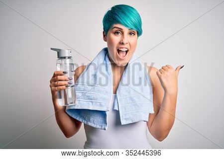 Young sporty woman with blue fashion hair holding bottle of water wearing towel after do sport pointing and showing with thumb up to the side with happy face smiling