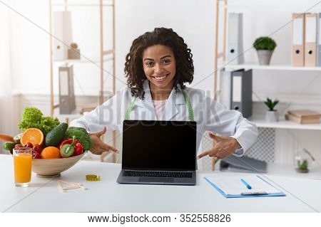 Cheerful Afro Woman Dietologist Pointing At Empty Laptop Screen, Sitting At Her Cabinet, Nutritionis