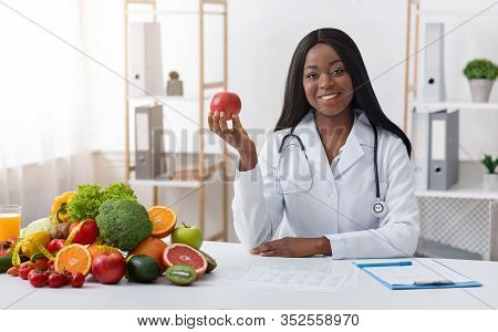 Beautiful Black Lady Dietologist Holding Apple In Her Hand, Recommending Fresh Fruits, Posing At Cli
