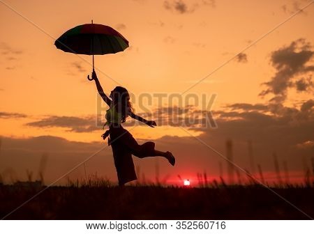 Silhouette Of Carefree Female With Umbrella Dancing In Field Against Cloudy Sunset Sky In Peaceful E