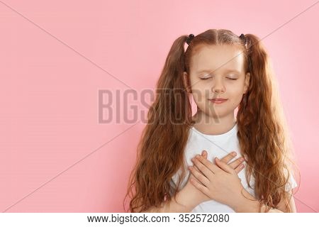 Cute Grateful Little Girl With Hands On Chest Against Pink Background. Space For Text