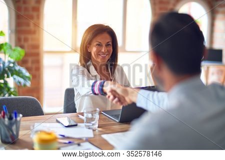 Two middle age business workers smiling happy and confident. Working together with smile on face using laptop doing handsake at the office