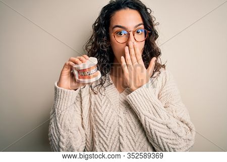 Young beautiful woman with curly hair holding plastic teeth with dental baces cover mouth with hand shocked with shame for mistake, expression of fear, scared in silence, secret concept