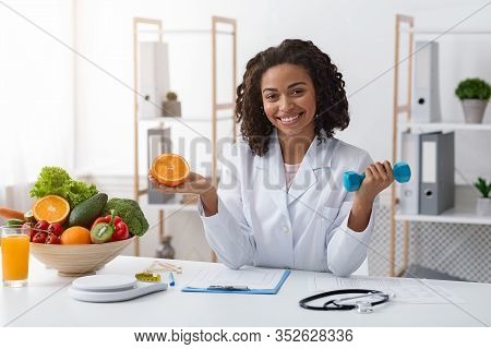 Young Cheerful Afro Woman Dietologist Holding Orange Half And Dumbbell In Her Hands, Recommending He