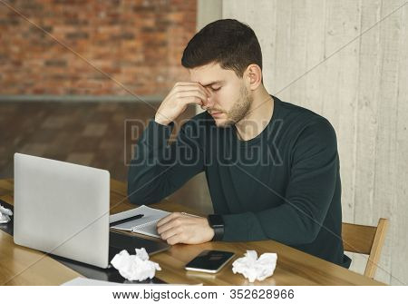 Tired Young Man Having Headache At Work, Touching Nosebridge Sitting At Laptop In Office