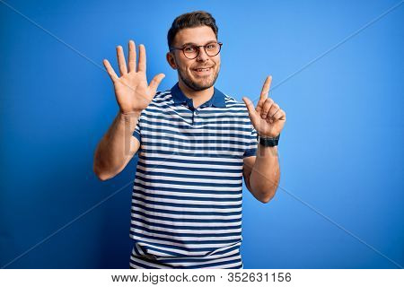 Young man with blue eyes wearing glasses and casual striped t-shirt over blue background showing and pointing up with fingers number seven while smiling confident and happy.