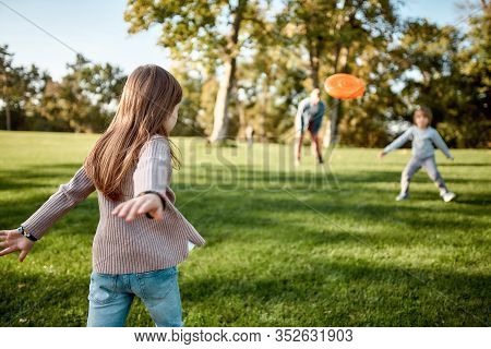Portrait Of Happy Family Of Four Playing With Disc On A Green Meadow With Grass. Focus On Daughter. 