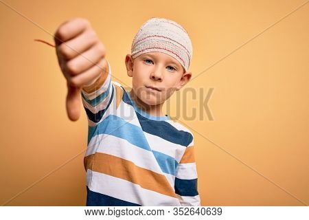 Young little caucasian kid injured wearing medical bandage on head over yellow background looking unhappy and angry showing rejection and negative with thumbs down gesture. Bad expression.