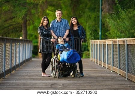 Caucasian Father Standing On Wooden Bridge At Park Outdoors With Biracial Children, One With Special