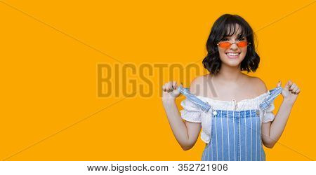 Smiling Caucasian Girl With Black Curly Hair And Eyewear Is Pulling Her Rompers On A Yellow Backgrou