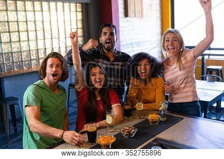 Front view of a group of five multi-ethnic male and female friends at the bar in a pub with arms in the air, cheering, having fun and celebrating while watching a sports game on TV, smiling and