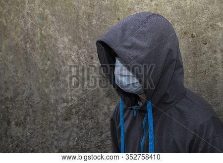 A Young Guy In A Motorcycle With A Hood In A Medical Mask On The Background Of A Concrete Wall. Pers