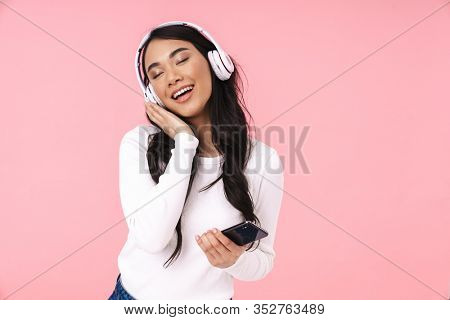 Cheerful pretty young asian woman listening to music with wireless headphones isolated over pink background, holding mobile phone