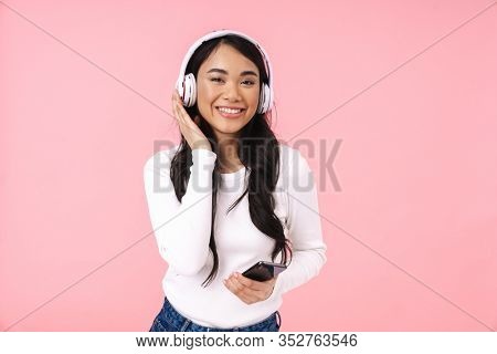 Cheerful pretty young asian woman listening to music with wireless headphones isolated over pink background, holding mobile phone